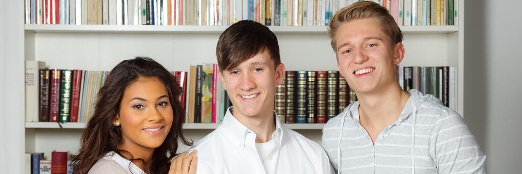 Drei Schülerinnen und Schüler lächeln vor einem Bücherregal in der Schulbibliothek in die Kamera.