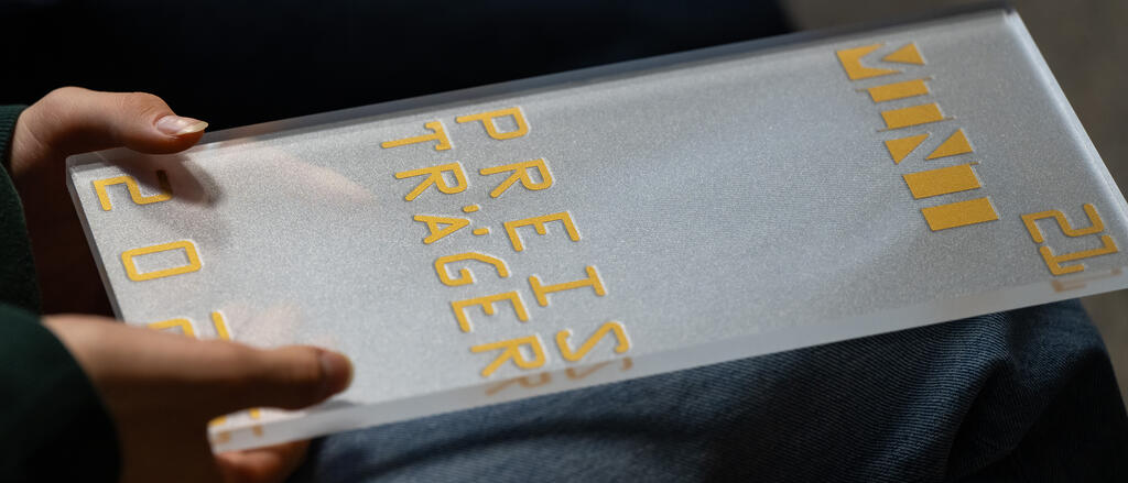 Zwei Hände sind zu sehen, die den Preispokal halten. Darauf steht "Preisträger 2025".