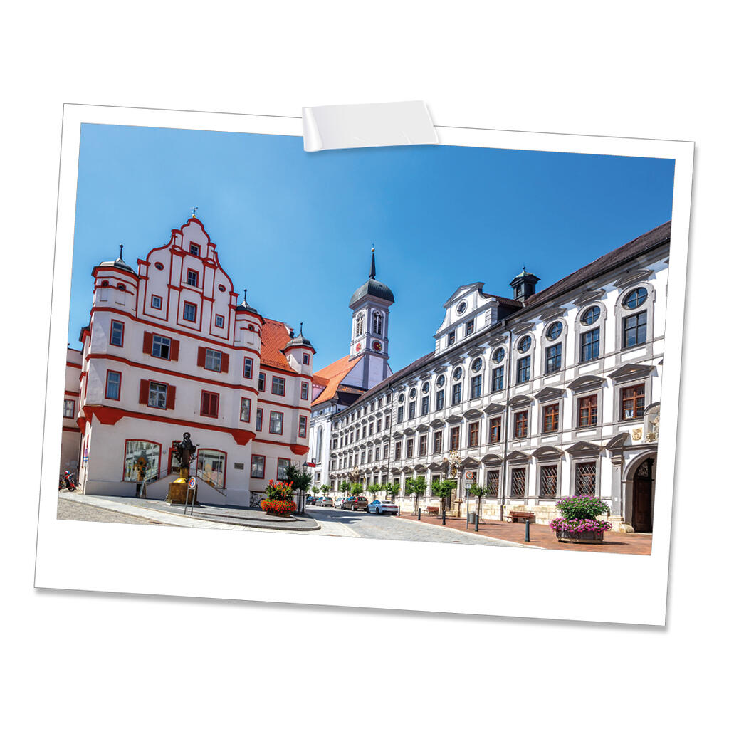 Die Aufnahme zeigt einen sonnigen Marktplatz mit historischen Gebäuden wie etwa einem weiß-roten Haus mit verzierten Giebeln. Im Hintergrund ragt ein Kirchturm mit grünem Zwiebeldach in den blauen Himmel.