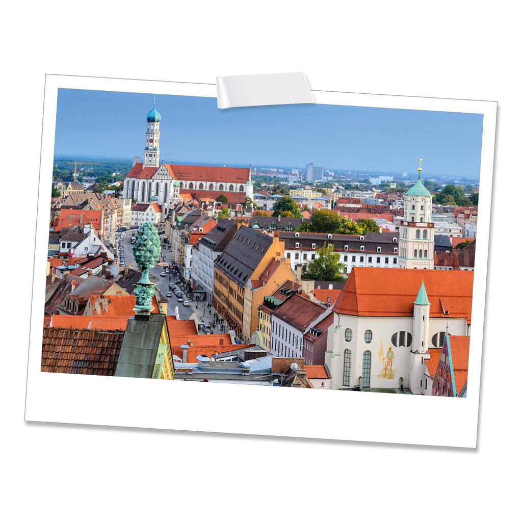 Die Aufnahme zeigt eine bayerische Stadt mit roten Dächern, historischen Gebäuden und zwei markanten Kirchtürmen unter blauem Himmel. Eine lange Straße zieht sich durch das Stadtzentrum.